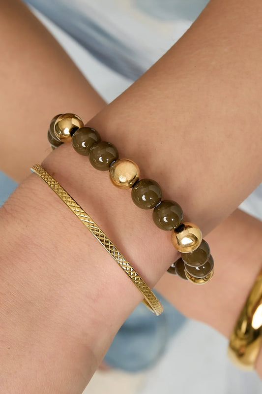 Model draagt gestapelde armbanden aan de pols waaronder bruine kralenarmband met gouden ballen, gecombineerd met gouden bangle en schakelarmband